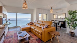 Ein modernes Wohnzimmer mit einer Ledersofa, einem Couchtisch und einem Essbereich mit einem schwarzen Tisch und Stühlen. Ein großes Fenster bietet Blick auf das Meer.