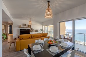 Ein modernes Wohnzimmer mit einer Couch, einem Esstisch gedeckt mit Tellern und Gläsern und einem großen Fenster mit Blick auf das Meer.