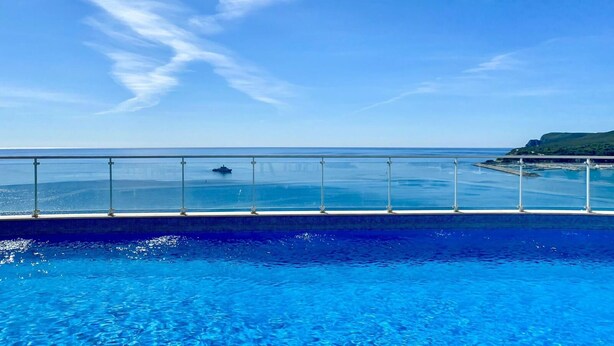 Ein klarer, blauer Himmel, ein Schwimmbecken mit Glasbarriere und ein Blick auf das Meer und einen grünen Hügel in der Ferne.