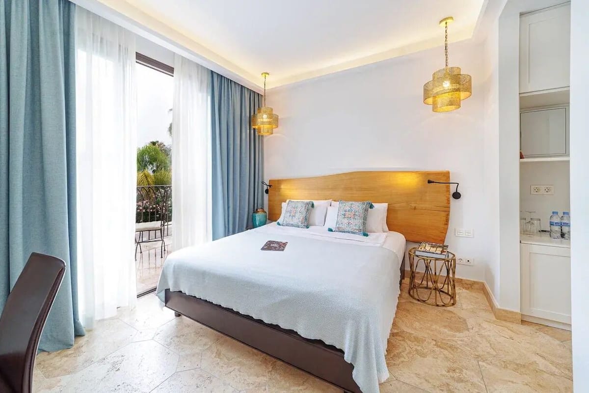 Quarto de hotel com uma cama grande, cabeceira de madeira, duas luminárias de cabeceira, uma mesinha e vista para o exterior.