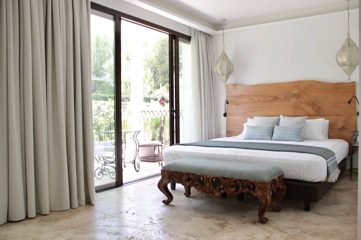 Quarto com uma cama grande, cabeceira de madeira, um banco e vista para uma varanda com mobiliário exterior.