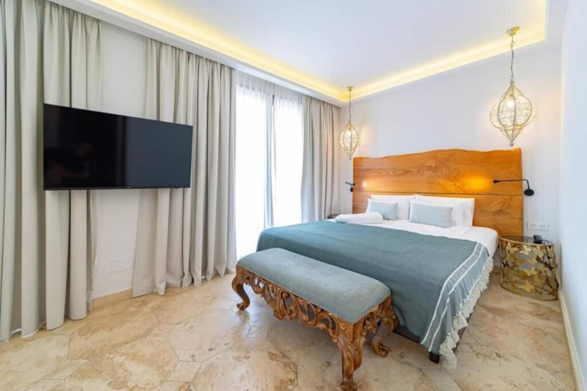 Quarto de hotel moderno com uma cama grande, televisão de tela plana e cabeceira de madeira.