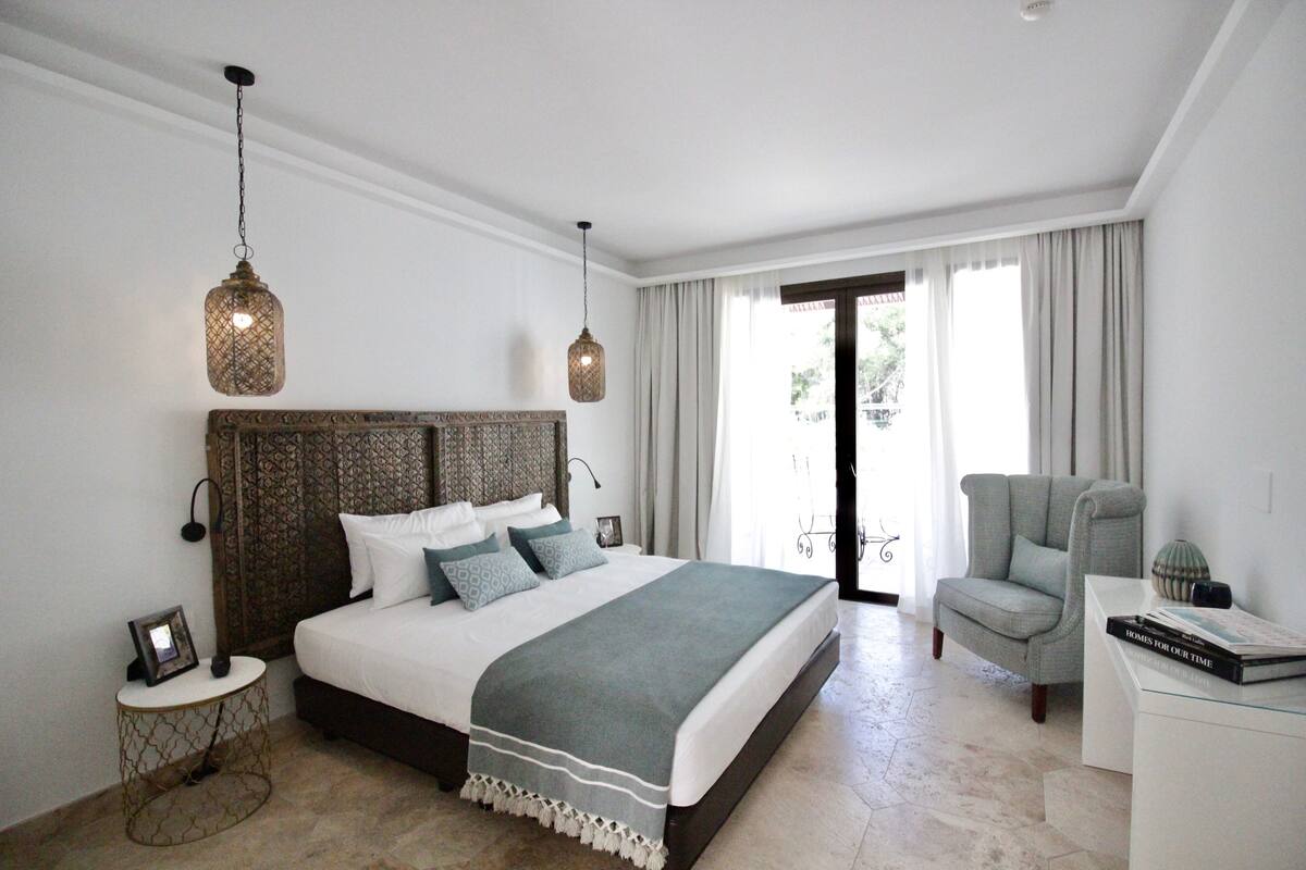Quarto com uma cama grande, uma cadeira, uma mesa de cabeceira e vista para o exterior.