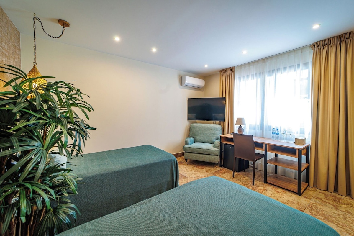 Quarto de hotel com duas camas, uma escrivaninha, uma cadeira, televisão e um vaso de planta.