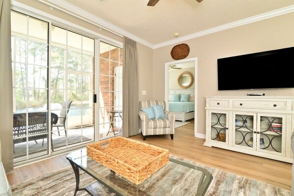 A spacious living room with a glass coffee table, a television mounted on the wall, and a sliding glass door leading to an outdoor patio.