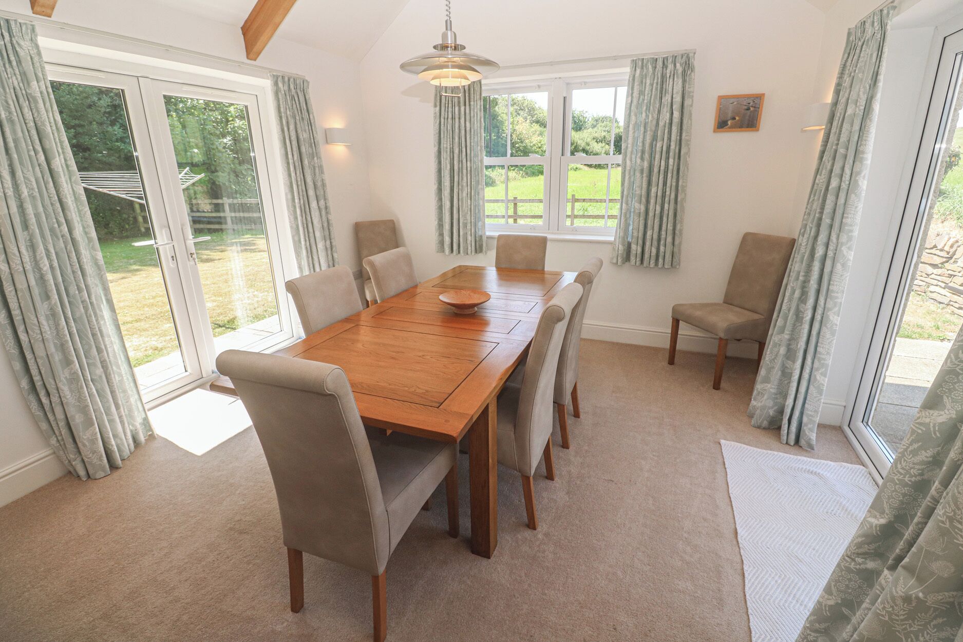 A dining room with a wooden table and chairs, large windows with curtains, and a hanging light fixture.