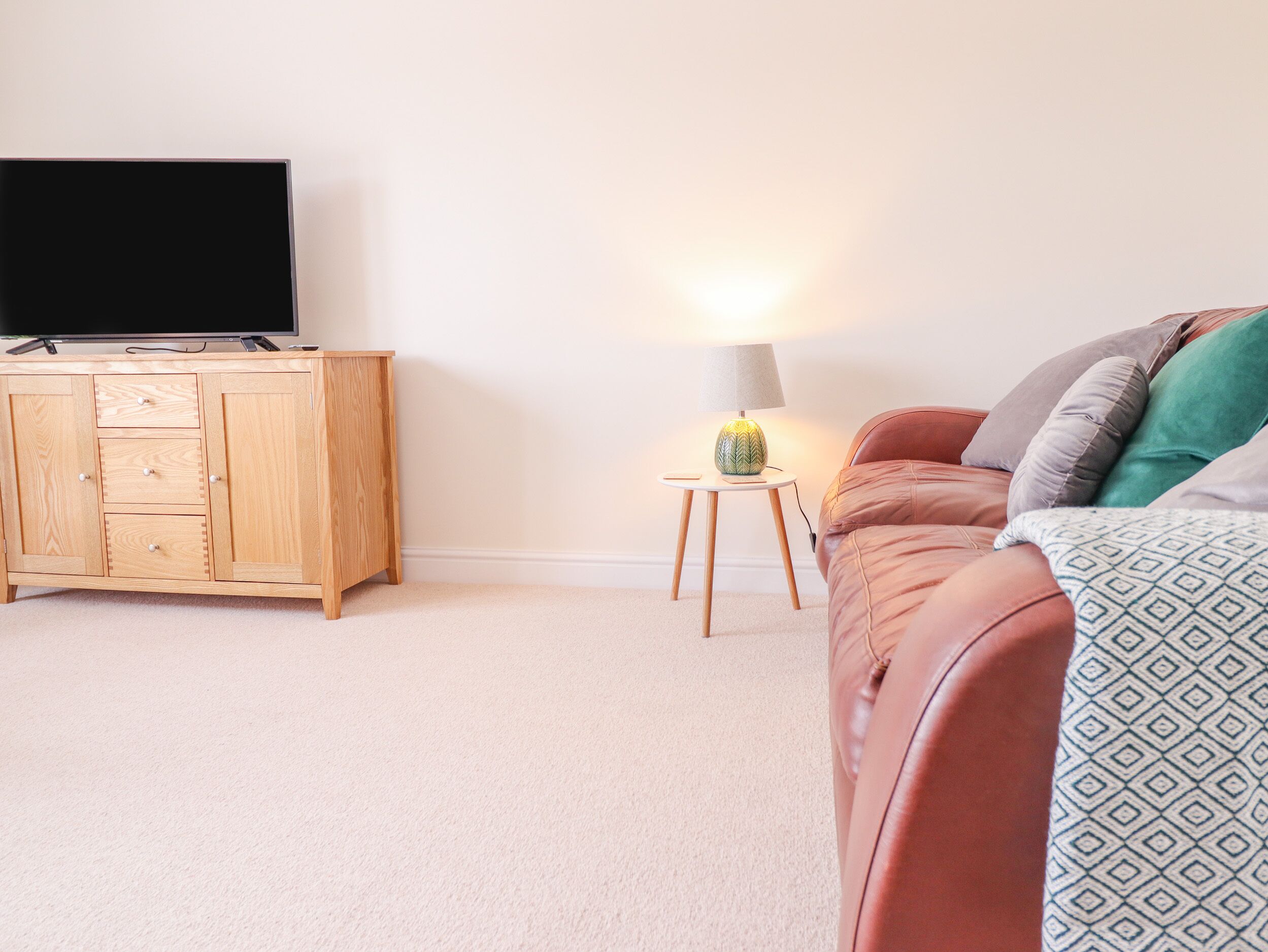 Ein Wohnzimmer mit einem Holzkasten, einem Fernseher darauf, einem kleinen Tisch mit einer Lampe und einem Ledersofa mit gemusterten Kissen.
