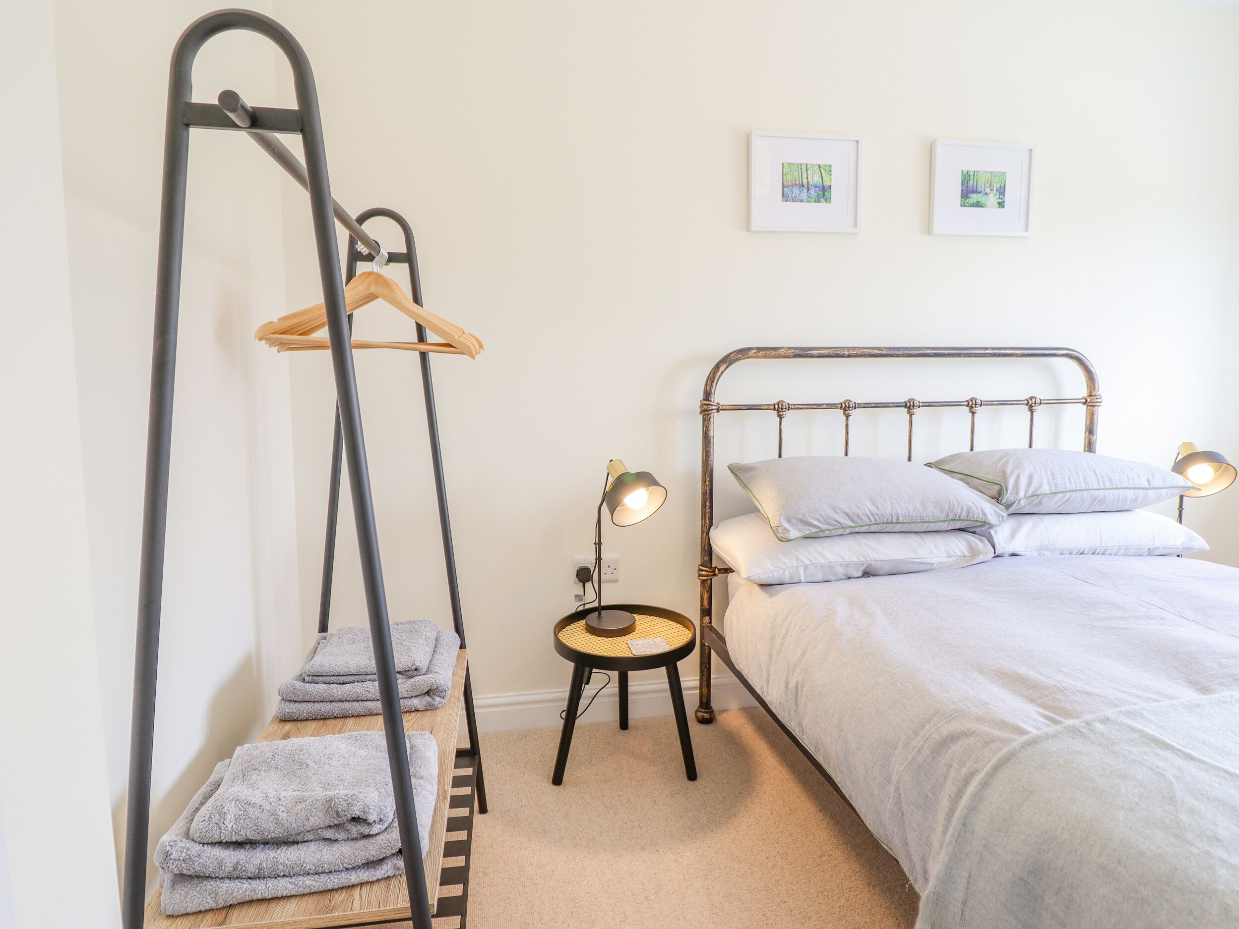 A bedroom with a metal bed frame, two bedside lamps, a small round table, a wooden drying rack, and two framed pictures on the wall.