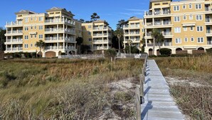 A beachfront residential building with multiple balconies, a boardwalk leading to the entrance, and a clear blue sky.