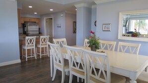 A dining area with a white table and chairs, a kitchen in the background, and a window with a view.