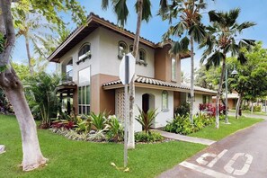 A two-story house with a tiled roof, surrounded by lush greenery and palm trees.