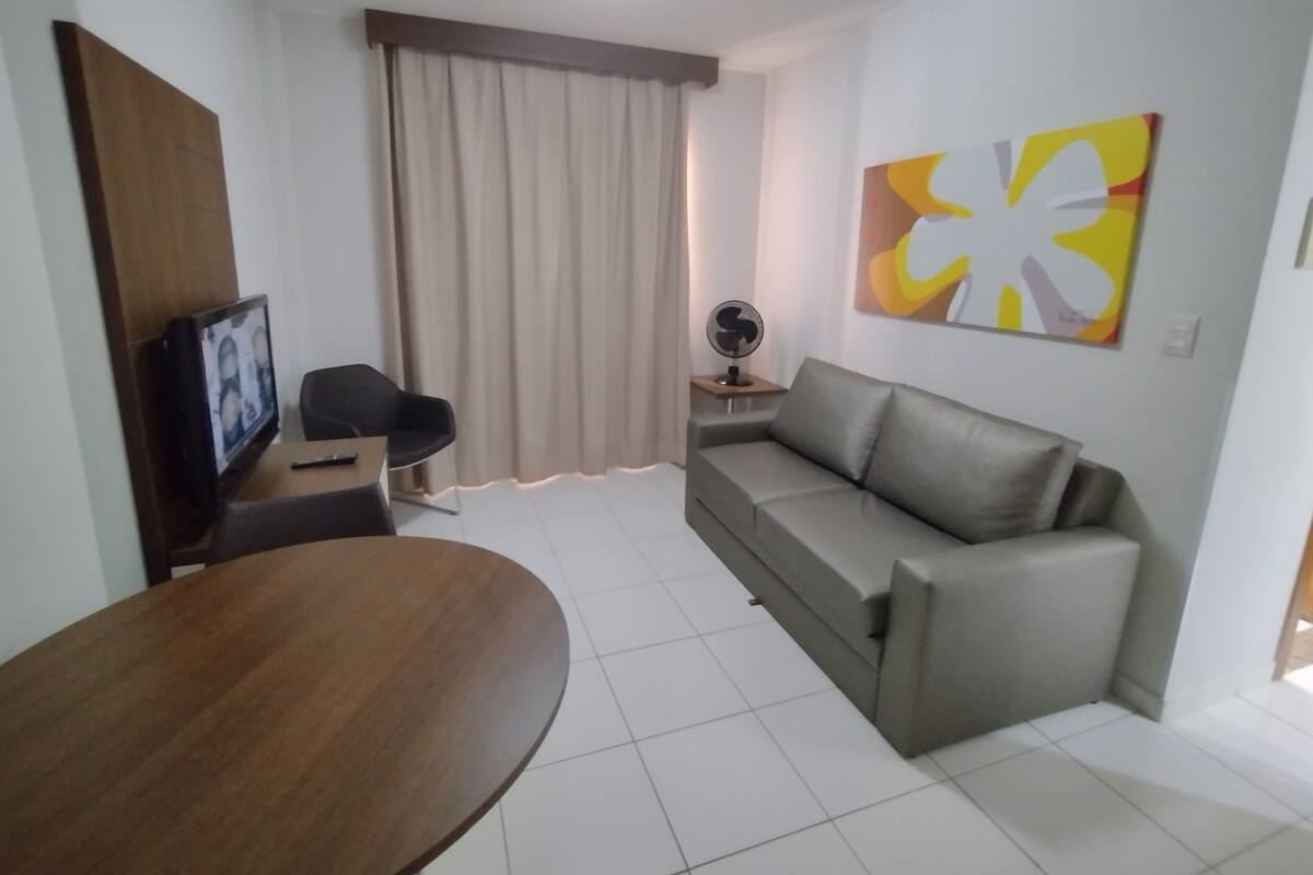 A modern living room with a grey sofa, a wooden dining table, a TV on a stand, a fan, and a large abstract painting on the wall.