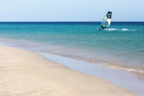 En strand med krystalklart turkis vand og en person som sejler på et windsurfbræt.