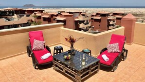 Una terraza en la azotea con dos sillas rojas acolchadas, una mesa de centro de vidrio y vista a la playa y edificios al fondo.