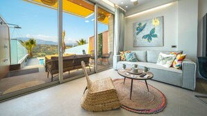 A modern living room with a sofa, a coffee table, and a wicker chair. There is a large window offering a view of the outdoors. - Oleas Suite Earth & Private Pool (Heraklion)