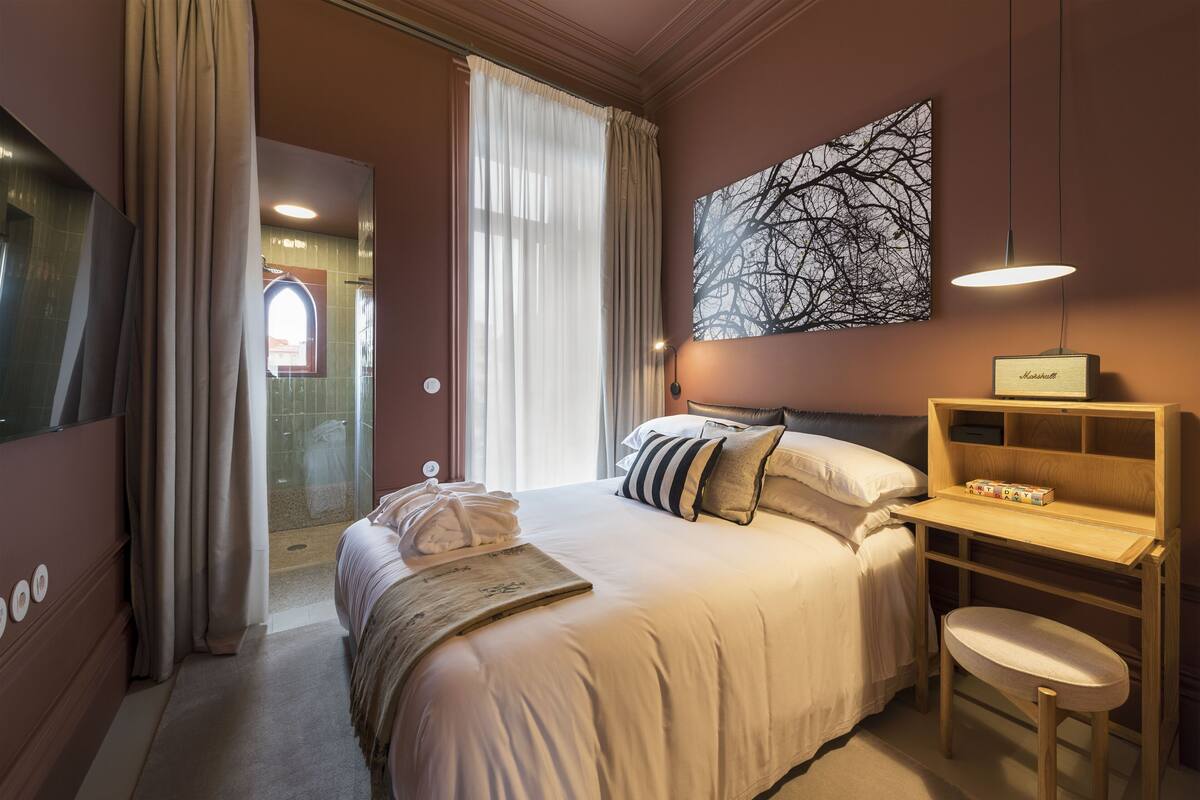A hotel room with a bed, a desk, a chair, a large window with curtains, a wall-mounted lamp, and a framed picture.