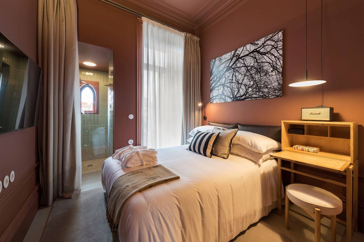 A hotel room with a bed, a desk, a chair, a large window with curtains, a wall-mounted lamp, and a framed picture.