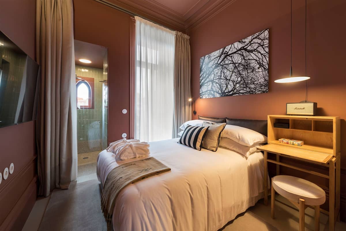 A hotel room with a bed, a desk, a chair, a large window with curtains, a wall-mounted lamp, and a framed picture.
