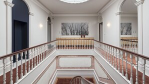 Una imponente escalera con balaustradas blancas y barandilla de madera, que conduce a un rellano con un amplio ventanal y un cuadro enmarcado de árboles desnudos.