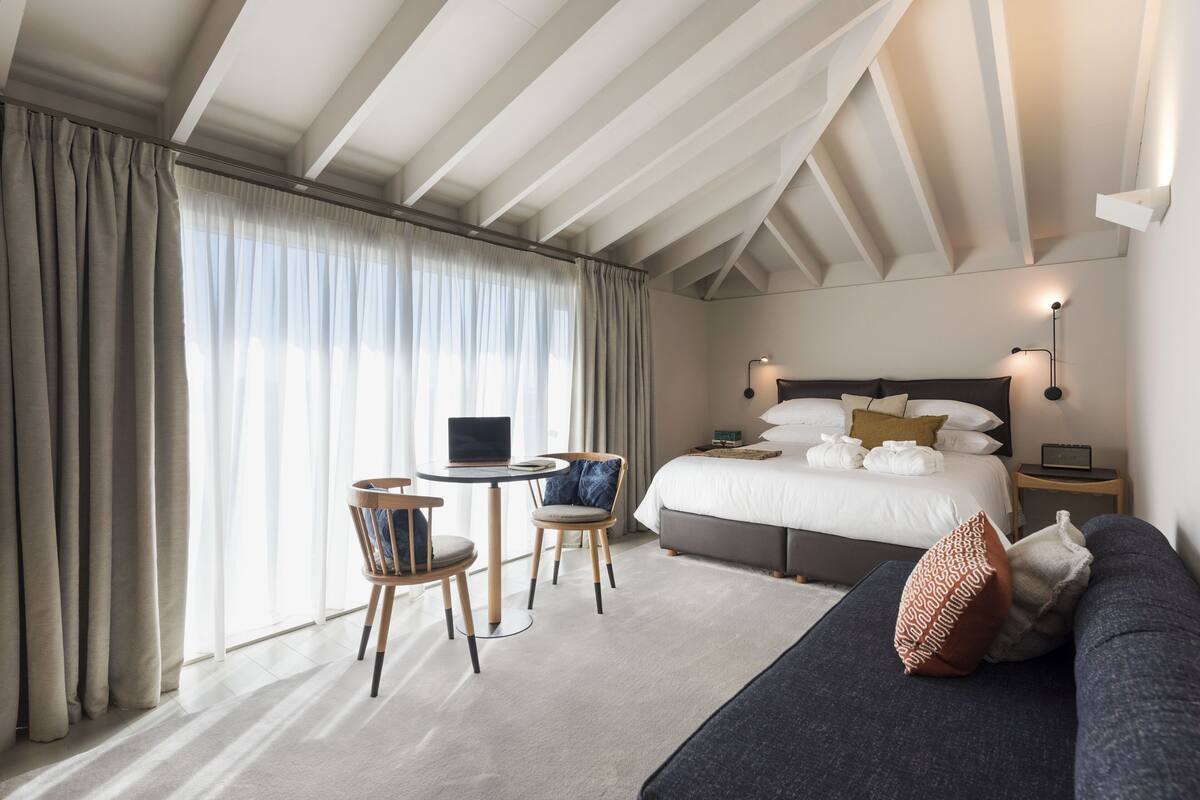 A modern hotel room with a large bed, a desk with a laptop, two chairs, and a sofa.