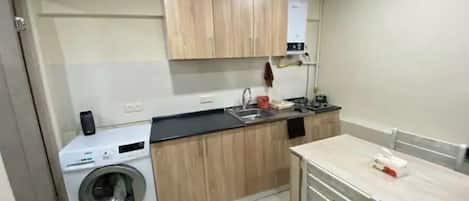 A compact kitchen with wooden cabinets, a built-in washing machine, and a dining area with a table and chairs.