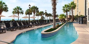 A pool area with lounge chairs and umbrellas, surrounded by palm trees and a view of the ocean.