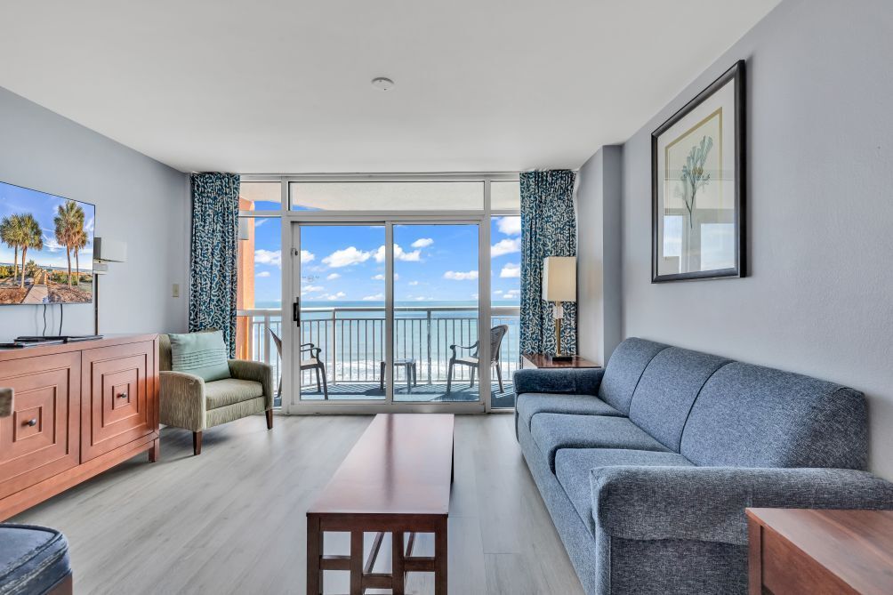 A modern living room with a sofa, a coffee table, a TV, and a balcony with a view of the ocean.