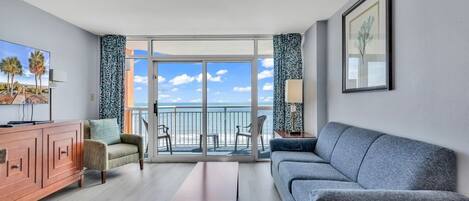A modern living room with a sofa, a coffee table, a TV, and a balcony with a view of the ocean.