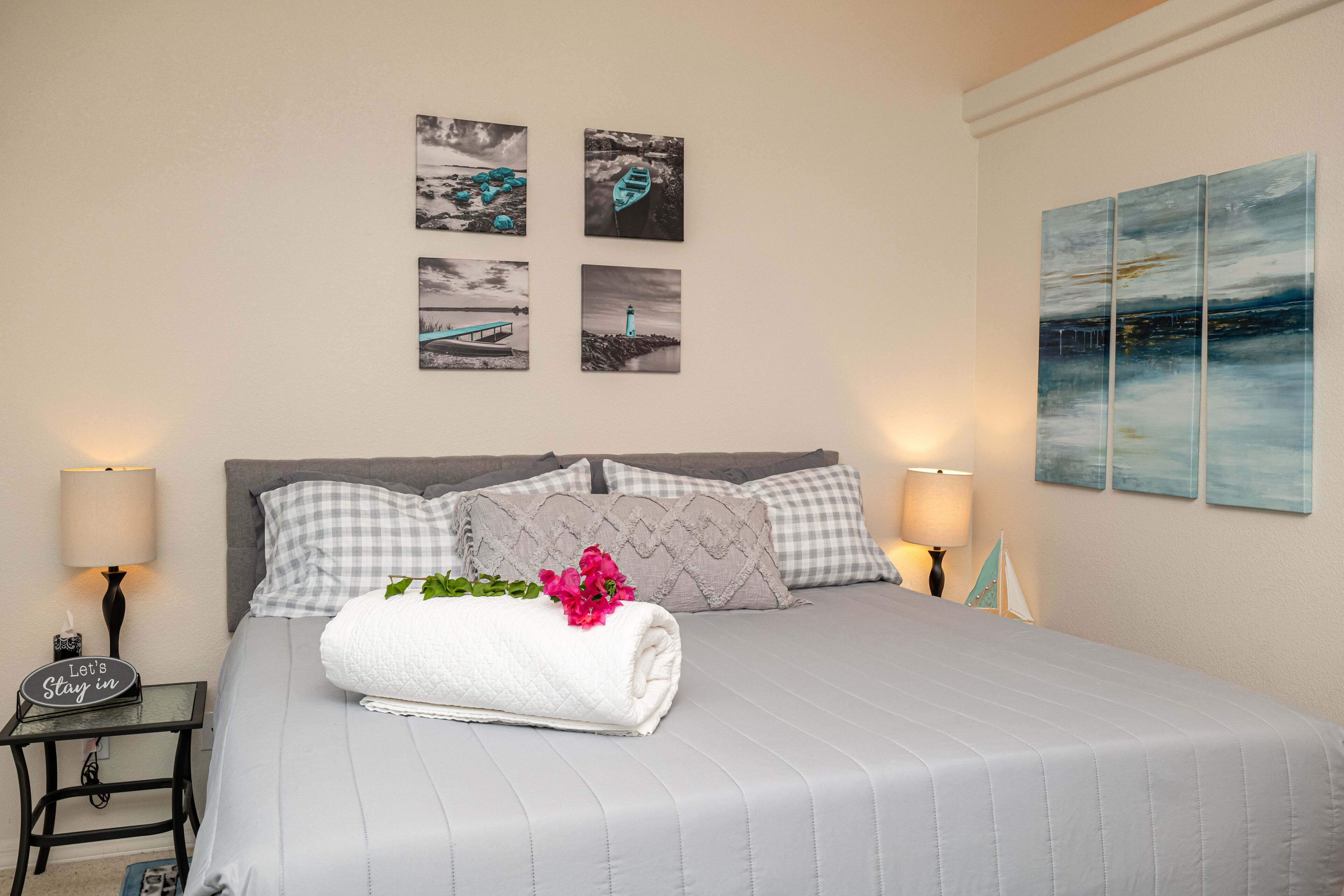 Una cama bien hecha con una toalla blanca enrollada y flores rosadas, dos lámparas de noche y cuatro cuadros en la pared.