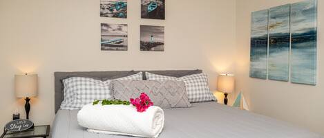 Una cama bien hecha con una toalla blanca enrollada y flores rosadas, dos lámparas de noche y cuatro cuadros en la pared.