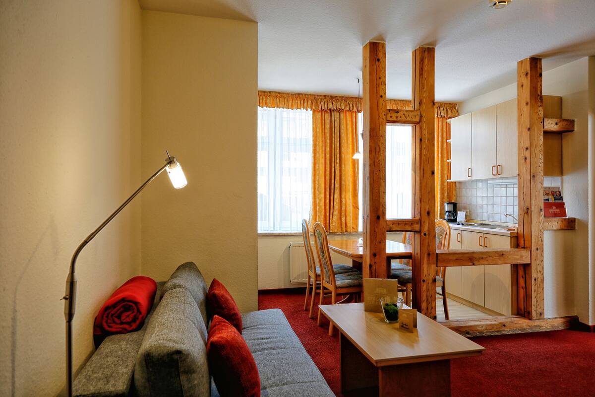 A compact living space with a sofa, a table, a lamp, and a kitchen area visible through an open doorway.