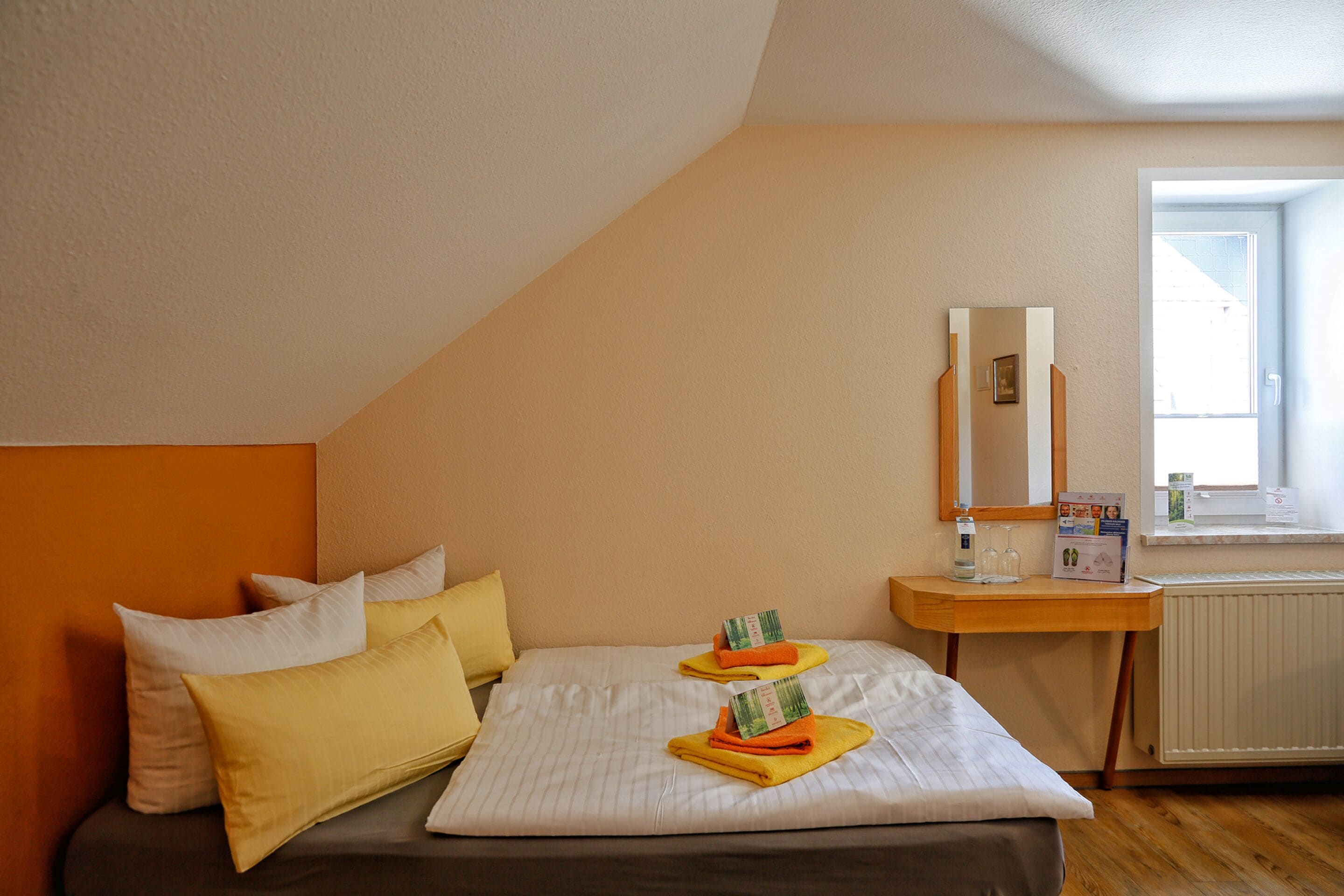 A bedroom with a bed, pillows, a bedside table with a mirror, a window, and a radiator.