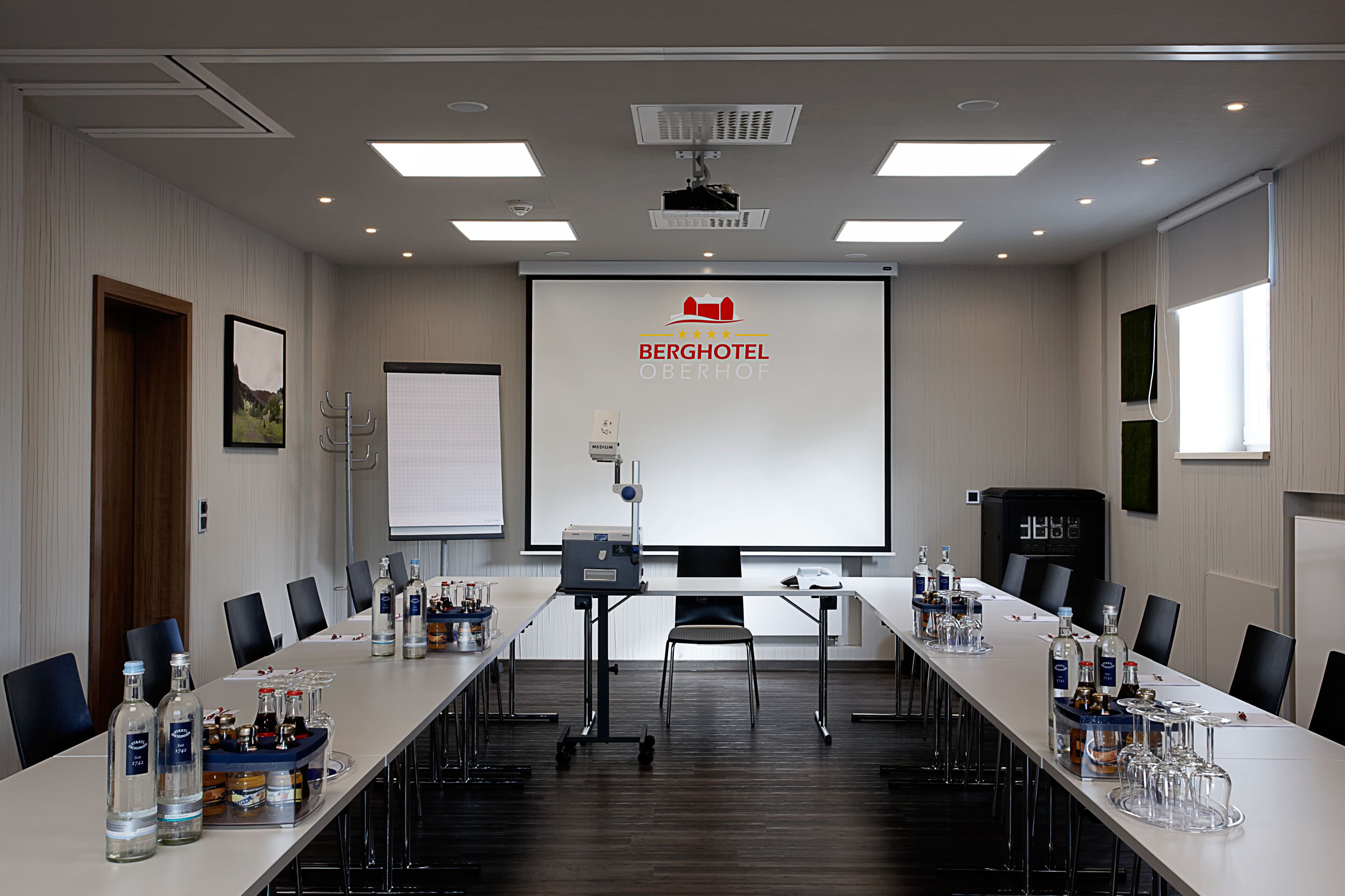 A conference room with a long table, chairs, a projector screen, and a presentation screen displaying the hotel name.