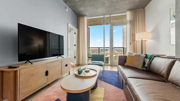 A modern hotel room with a large flat-screen TV, a wooden sideboard, a sofa, a coffee table, and a balcony with a view. - Laredo - Rooftop pool & Rainey St views (Austin)