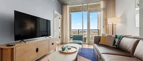 A modern hotel room with a large flat-screen TV, a wooden sideboard, a sofa, a coffee table, and a balcony with a view.