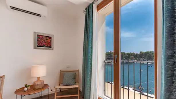 A room with a wooden floor, a chair, a table with a lamp, a framed picture on the wall, and a large window with a view of the sea and a balcony. - Mamma mia luxury rooms - Double Room with Sea View (Trogir)