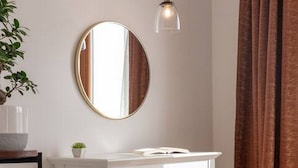 Une pièce avec un bureau blanc, un miroir rond, un luminaire suspendu, une plante en pot et un rideau marron.