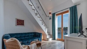 Een moderne woonkamer met een blauwe bank, een rieten stoel, een witte kast en een trap naar een balkon met uitzicht.