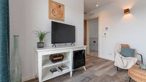 Een moderne woonkamer met een flatscreen televisie op een witte console, een potplant en een rieten bijzettafeltje met kussen.