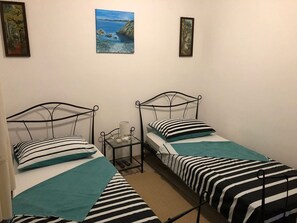 A room with two single beds, each with a metal frame and striped bedding. There is a small bedside table with a lamp and a remote control. The walls are plain, and there are two framed pictures above the beds. - Apartment Villa Jakov - Two Bedroom Apartment with Terrace and Swimming Pool (Rogač)