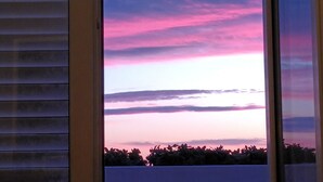 Une fenêtre donnant sur un ciel au coucher du soleil, un horizon lointain et la silhouette d’un feuillage.