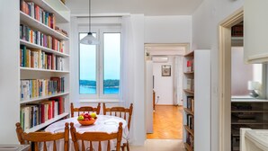 Un coin repas avec une table dressée pour quatre personnes, entouré d’étagères à livres, une vue sur la mer par la fenêtre, et un espace cuisine visible sur la droite.