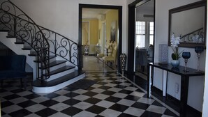 Un suelo a cuadros, una escalera con barandillas ornamentales, un comedor con sillas y una consola con adornos.