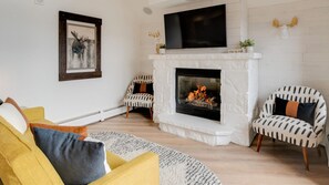 A living room with a white fireplace, two patterned chairs, a yellow sofa, and a framed deer artwork on the wall.