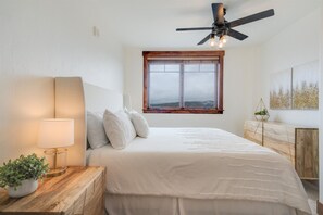 A bedroom with a bed, a nightstand with a lamp, a window with a view, and a ceiling fan.