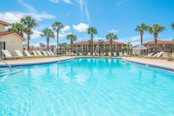 A swimming pool with lounge chairs and palm trees, surrounded by a building with a red roof.