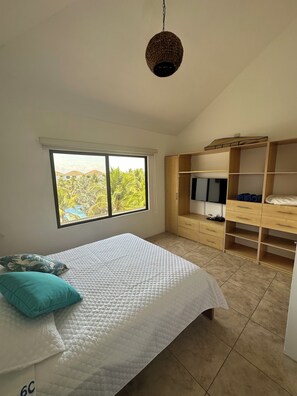 Ein Schlafzimmer mit Bett, Kleiderschrank, Fernseher und Fenster mit Blick auf Palmen.