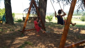 Zwei Kinder spielen auf einer Holzschaukel in einer grünen Wiese mit Bäumen und weiter Aussicht auf eine Stadt.