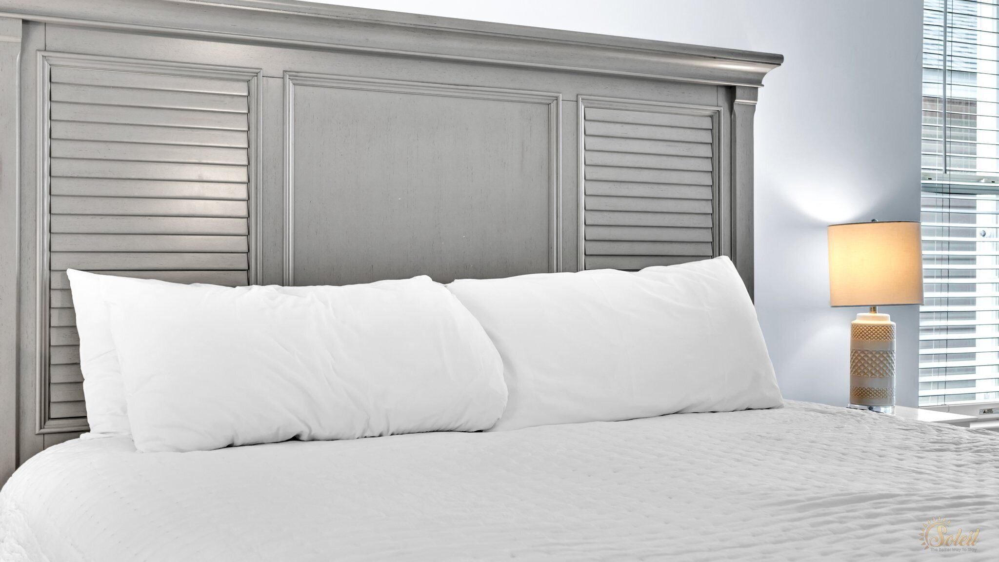A neatly made bed with white linens and pillows, a wooden headboard, and a bedside lamp.