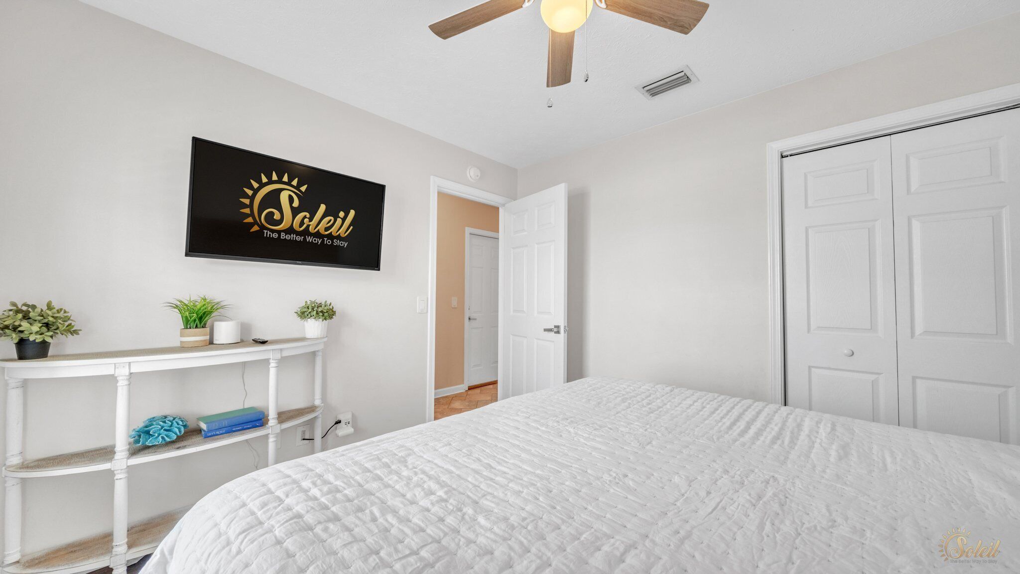 Un dormitorio con cama, ventilador de techo, un cartel en la pared, una puerta blanca y un pasillo que conduce a otra habitación.